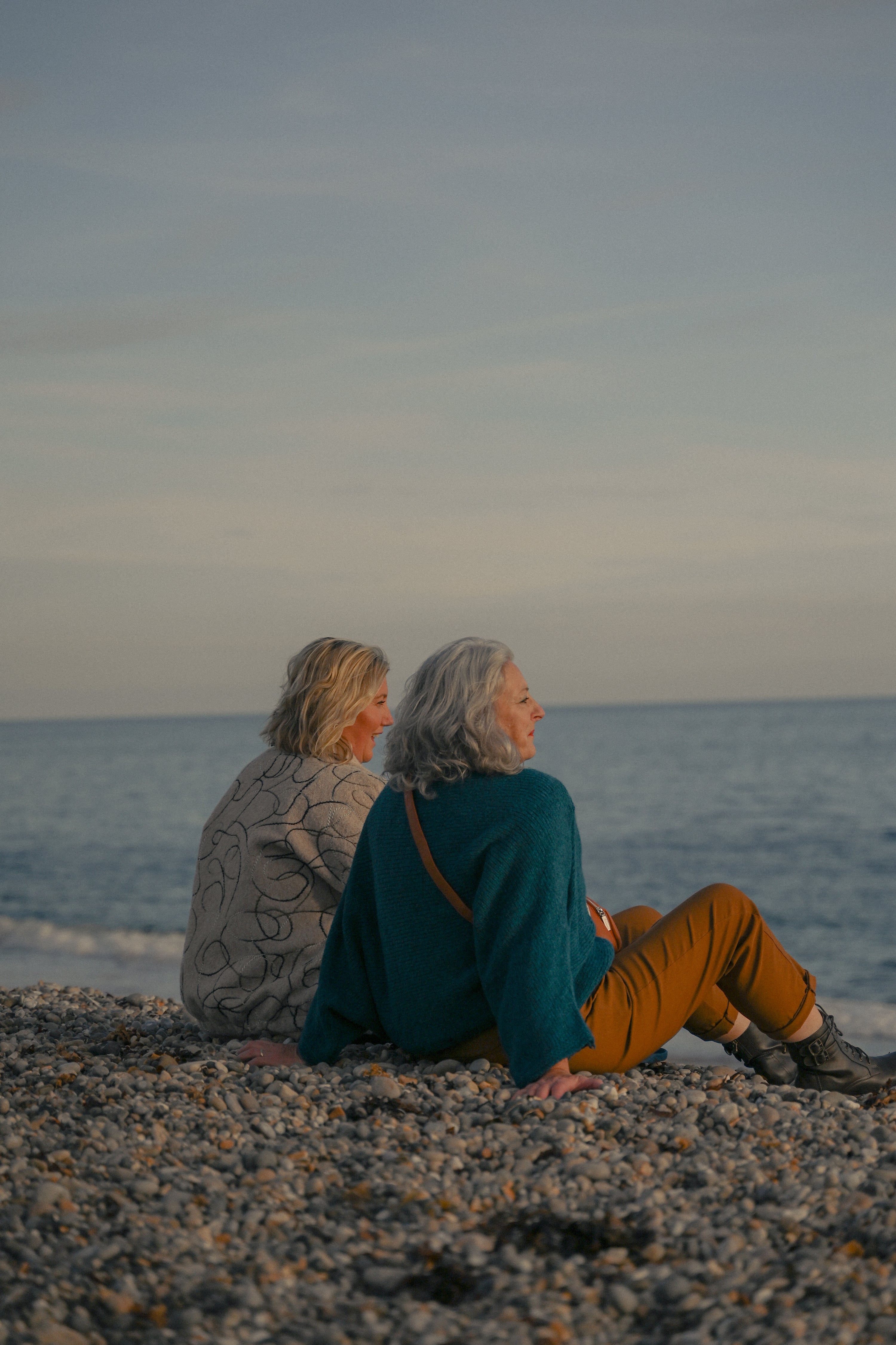 Two people sitting on a pebbly beach looking out at the ocean.